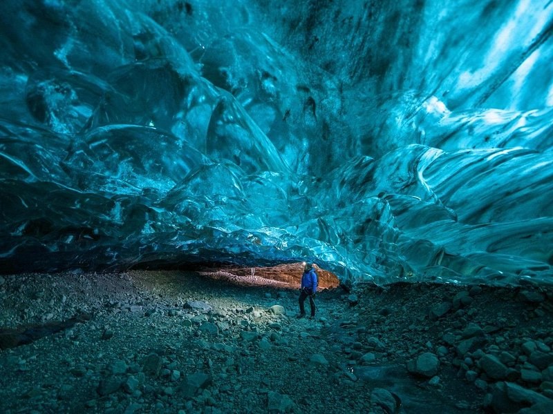 A vast turquoise ice cave roof over a gravel floor, one visitor small beneath it