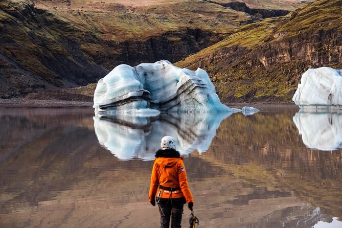 A hiker in crampons facing a banded iceberg stranded in a still glacier lagoon