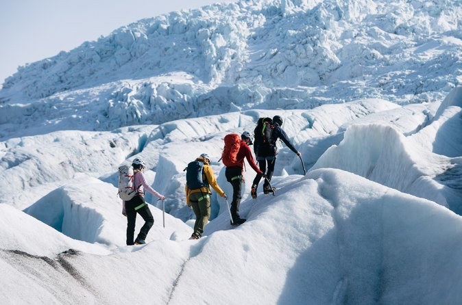 A roped line of hikers in crampons climbing between ice ridges below an icefall