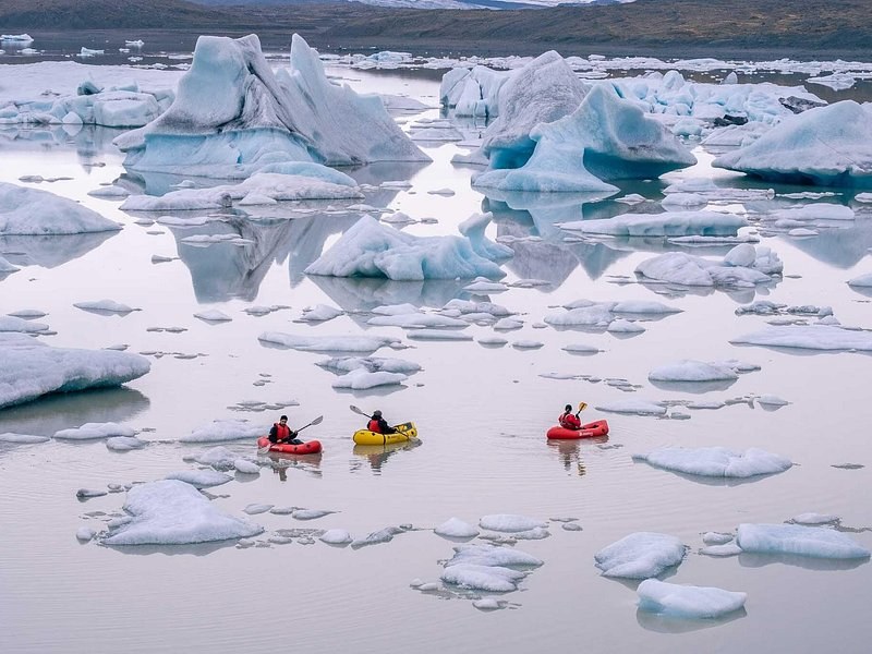 Three kayaks paddling between icebergs on a still glacier lagoon, seen from above