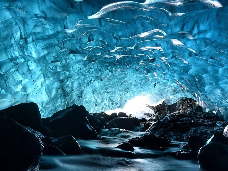 The rippled blue ceiling of an ice cave over black rocks and running meltwater