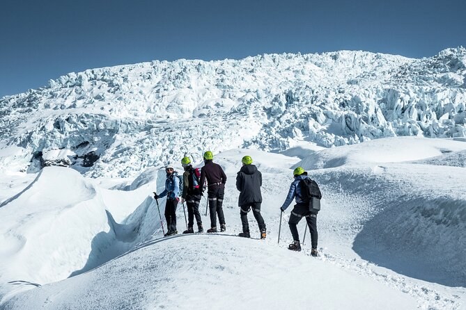 Five hikers with ice axes on white ice below a jagged icefall
