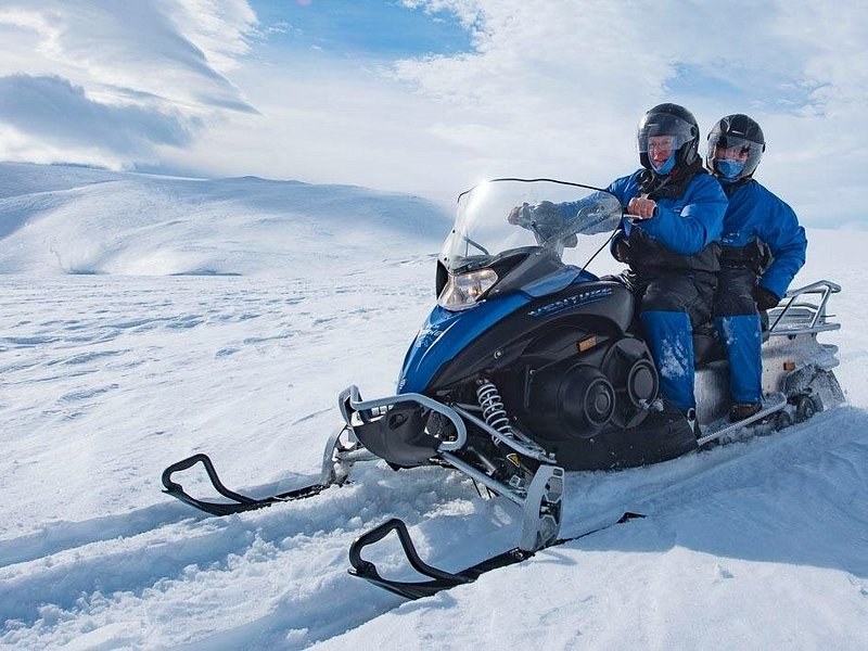 Two riders in helmets and blue overalls sharing one snowmobile on a snowfield