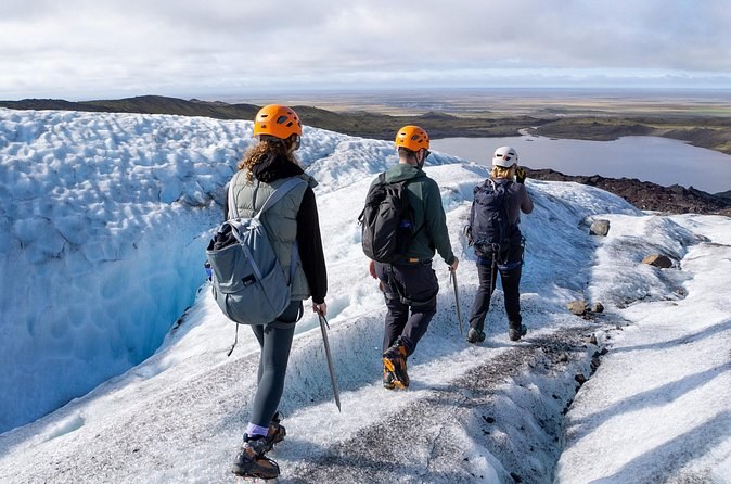 Three helmeted hikers walking down a glacier toward a lagoon and the flat plain